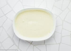A white oval dish filled with a smooth, creamy white sauce, perfect for serving Eggless Chicken Francese (Chicken French), sits on a white tiled surface.