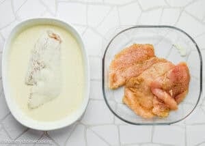 Raw, seasoned chicken breasts in a glass dish next to one chicken breast being coated in a creamy batter for Eggless Chicken Francese (Chicken French), on a white tiled surface.