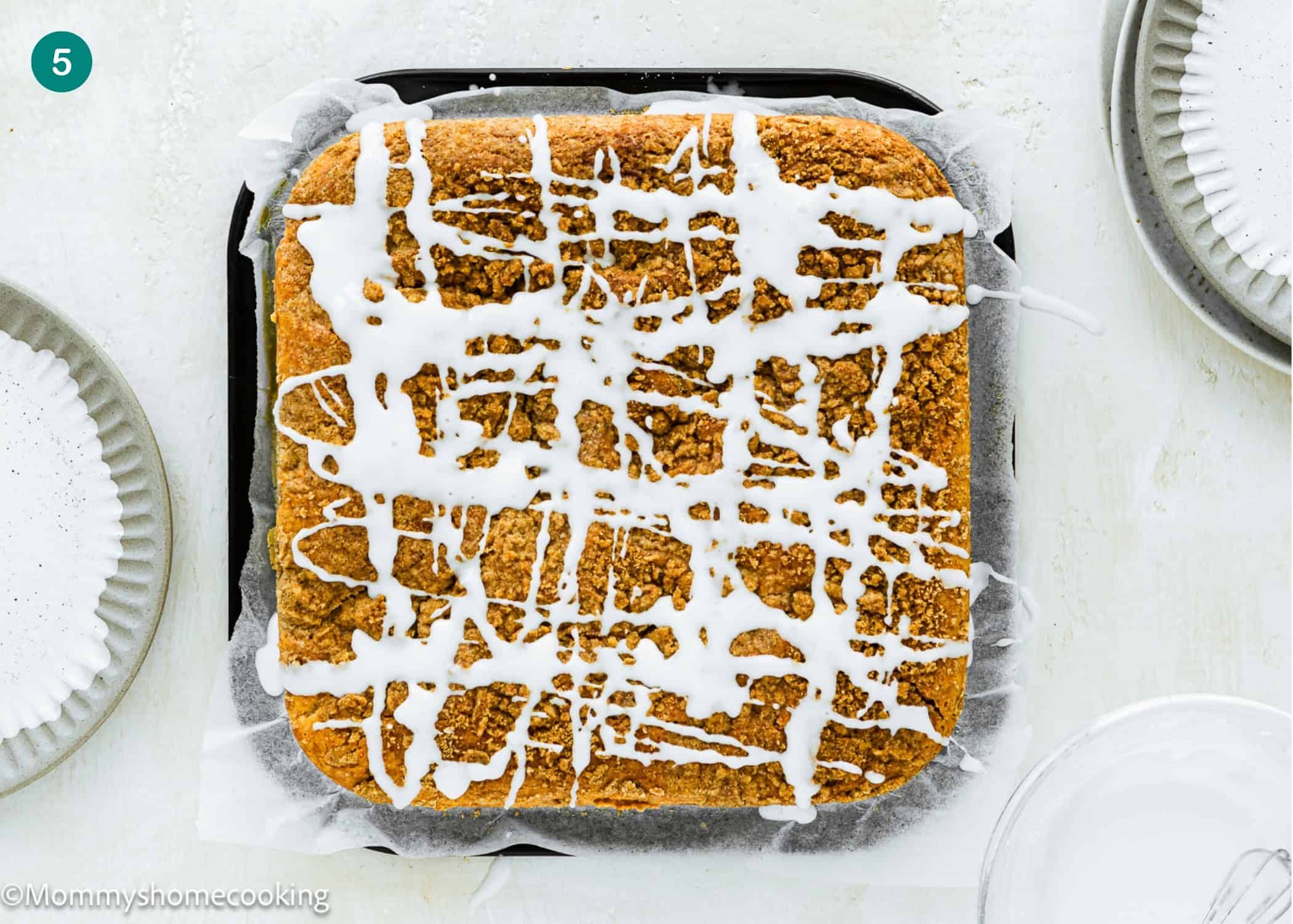 An Eggless Cream Cheese Coffee Cake with white icing drizzle sits on parchment paper in a baking pan, surrounded by three empty plates.