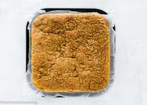 An Eggless Cream Cheese Coffee Cake with a crumbly streusel topping sits on parchment paper in a black baking pan against a white background.