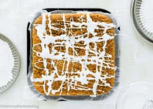 An Eggless Cream Cheese Coffee Cake topped with white icing drizzle sits on parchment paper in a baking pan, with plates and a bowl nearby on a light-colored surface.