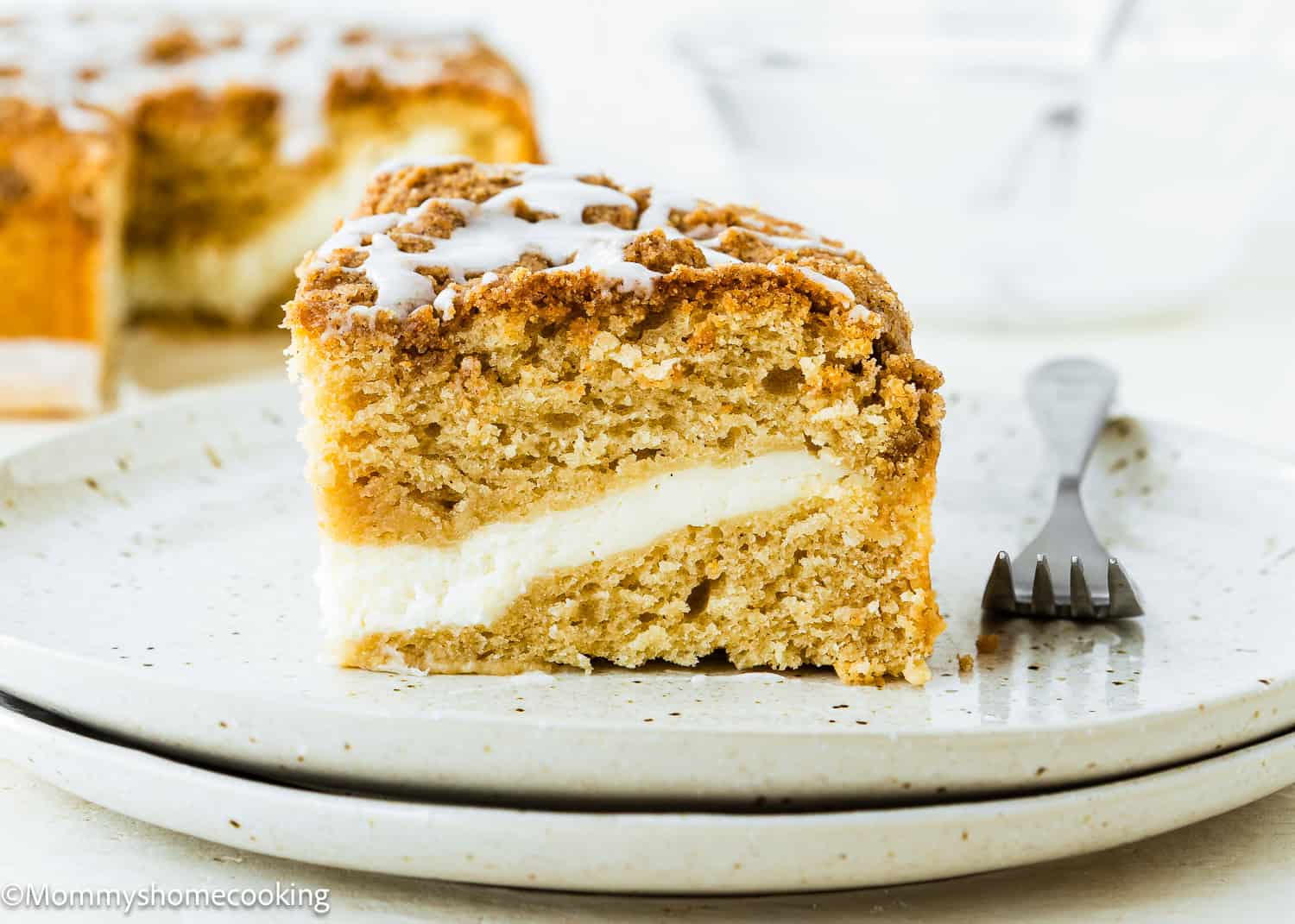 A slice of Eggless Cream Cheese Coffee Cake with a cream cheese filling, topped with icing, sits on a speckled plate with a fork beside it.