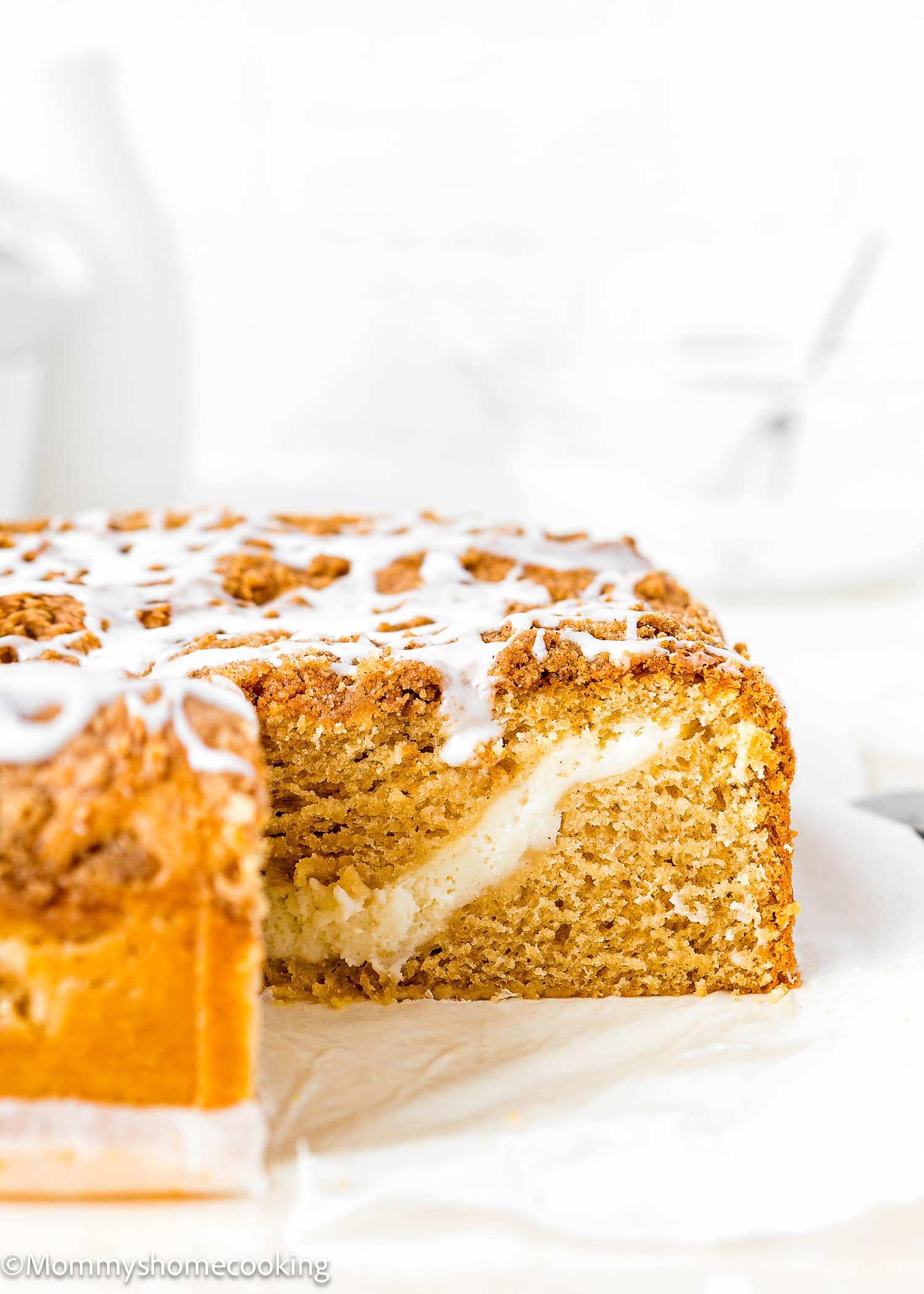 A close-up of an Eggless Cream Cheese Coffee Cake with a crumb topping and white icing, revealing a creamy filling inside.