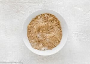 A white bowl filled with a light brown, crumbly Eggless Cream Cheese Coffee Cake mixture sits on a textured white surface.