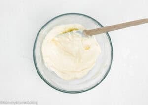 A glass bowl filled with creamy white mixture for an Eggless Cream Cheese Coffee Cake being stirred with a spatula on a light surface.