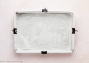 A rectangular baking pan lined with parchment paper and secured with four binder clips, ready for baking Eggless Ooey Gooey Butter Bars (Neiman Marcus Bars), sits on a light pink surface.