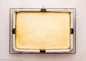 A large, rectangular Eggless Ooey Gooey Butter Bars (Neiman Marcus Bars) cheesecake sits in a parchment-lined pan on a cooling rack against a light background.
