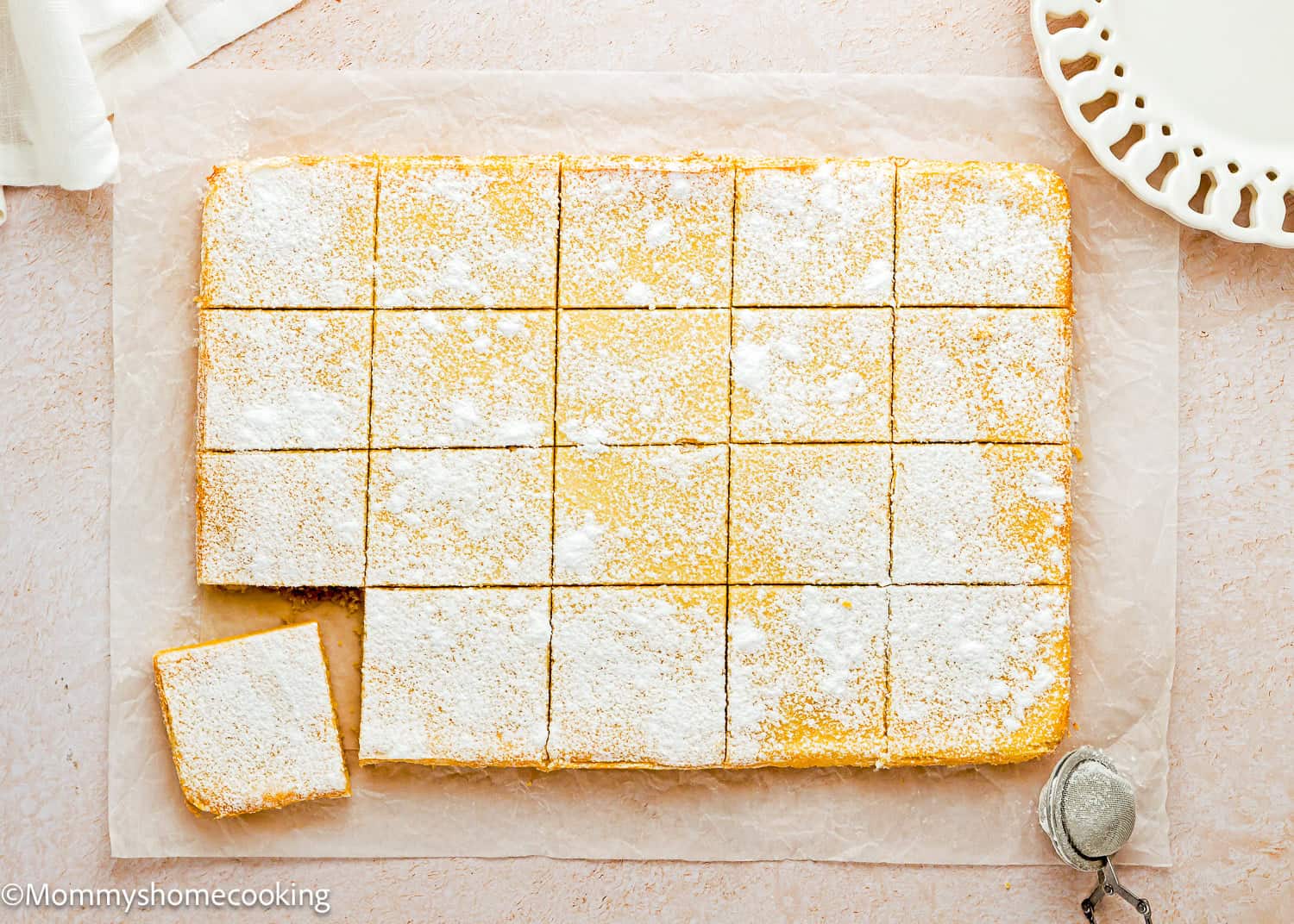 Rectangular lemon bars, inspired by Eggless Ooey Gooey Butter Bars (Neiman Marcus Bars), dusted with powdered sugar, cut into 20 squares on parchment paper—one square slightly pulled out with a white plate nearby.
