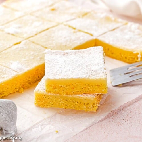 Two Eggless Ooey Gooey Butter Bars (Neiman Marcus Bars) stacked on parchment paper, sprinkled with powdered sugar, with more bars cut into squares in the background. A metal spatula and powdered sugar sifter are nearby.