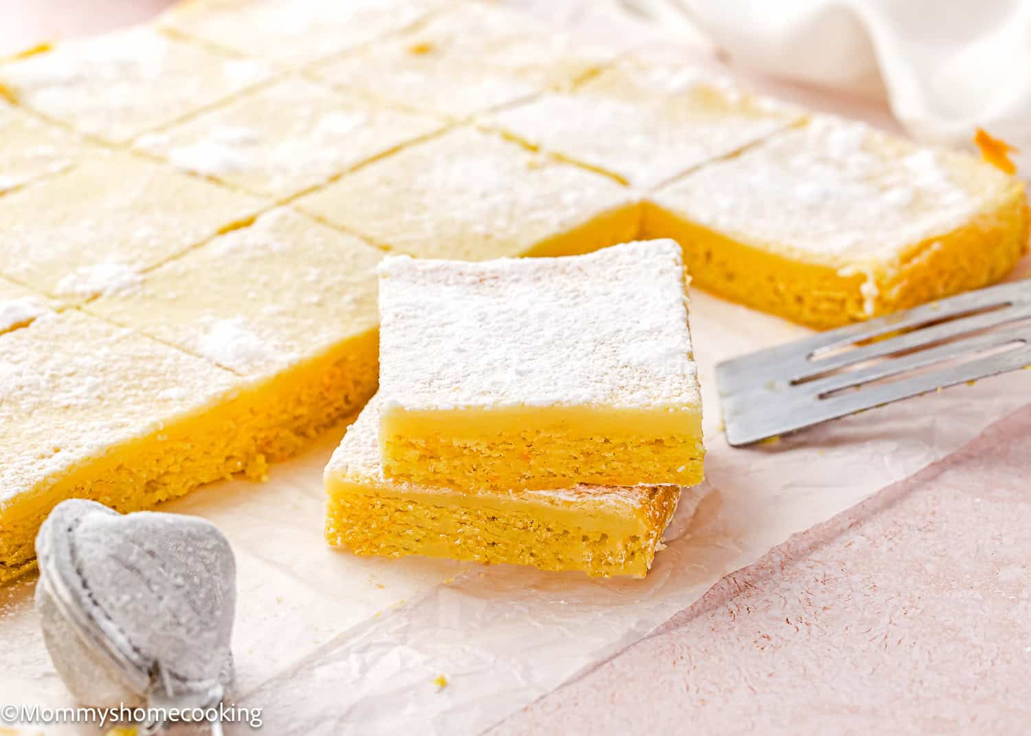 Two Eggless Ooey Gooey Butter Bars (Neiman Marcus Bars) stacked on parchment paper, sprinkled with powdered sugar, with more bars cut into squares in the background. A metal spatula and powdered sugar sifter are nearby.