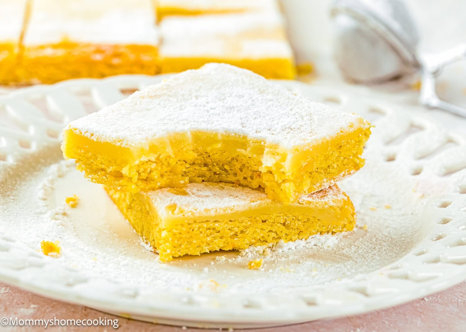Two Eggless Ooey Gooey Butter Bars (Neiman Marcus Bars) stacked on a white plate, dusted with powdered sugar. The top bar has a bite taken out of it. More lemon bars are visible in the background.