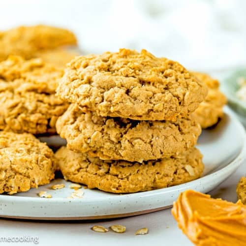 A plate of Eggless Peanut Butter Oatmeal Cookies stacked on top of each other, with a spoonful of peanut butter and oats visible nearby.