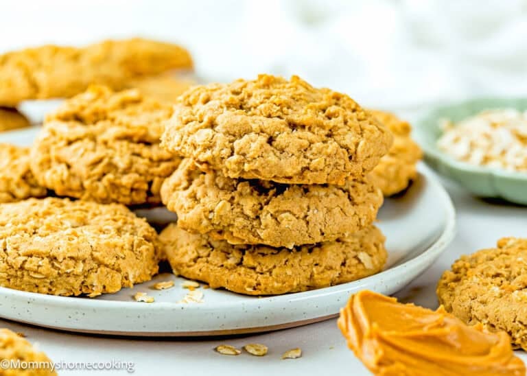 A plate of Eggless Peanut Butter Oatmeal Cookies stacked on top of each other, with a spoonful of peanut butter and oats visible nearby.