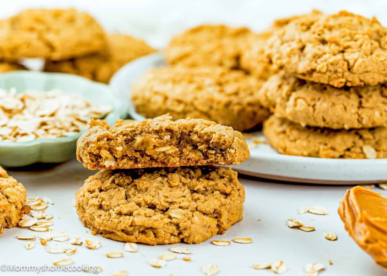 A stack of Eggless Peanut Butter Oatmeal Cookies is displayed on a plate, with one cookie broken in half in the foreground, surrounded by oats and a dollop of peanut butter.