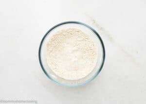 A glass bowl filled with flour sits on a white surface, ready to be used for making Eggless Peanut Butter Oatmeal Cookies.