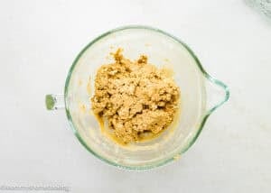 A glass mixing bowl containing a rough, beige Eggless Peanut Butter Oatmeal Cookies dough mixture sits on a white surface.