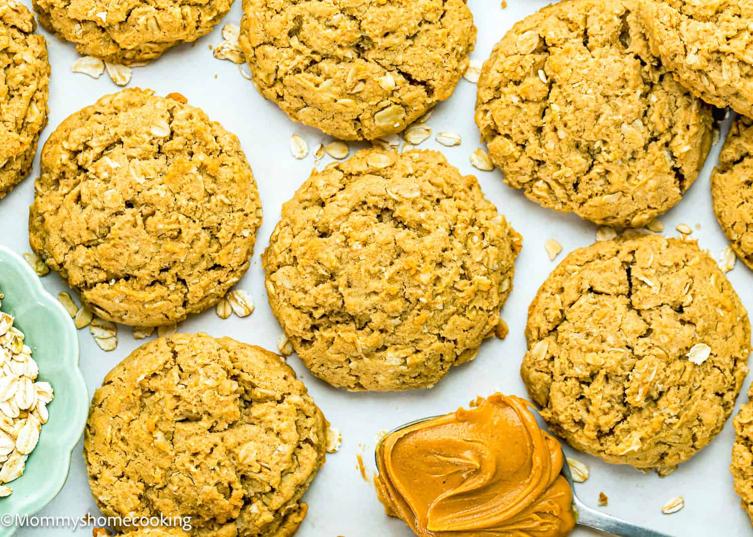 Several Eggless Peanut Butter Oatmeal Cookies are arranged on a white surface with a spoonful of peanut butter and some loose oats nearby.