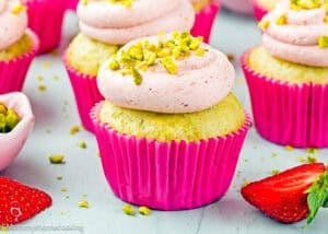 Eggless Pistachio Cupcakes with Strawberry Buttercream, topped with pink frosting and chopped pistachios in a pink wrapper, surrounded by strawberries and more cupcakes.
