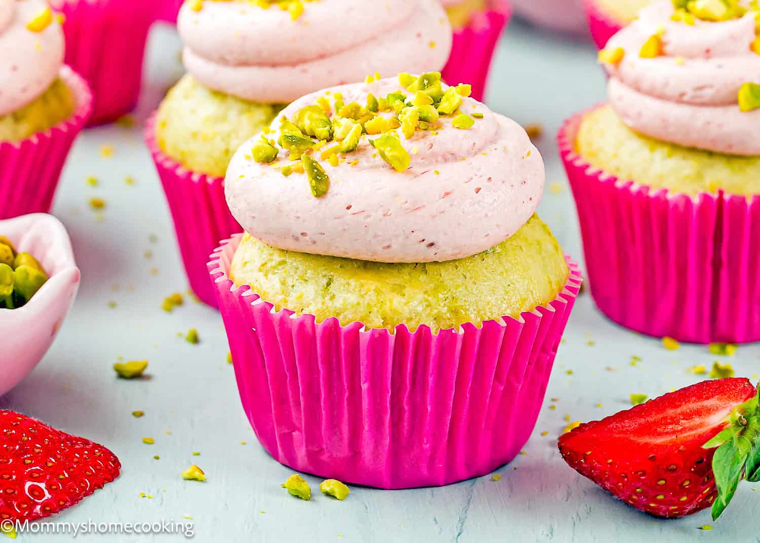 Eggless Pistachio Cupcakes with Strawberry Buttercream, topped with pink frosting and chopped pistachios in a pink wrapper, surrounded by strawberries and more cupcakes.