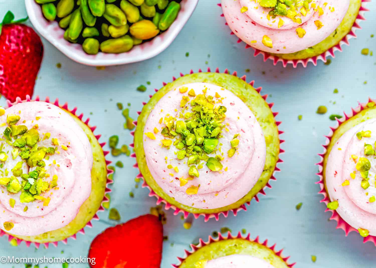 Top view of Eggless Pistachio Cupcakes with Strawberry Buttercream, topped with pink frosting and chopped pistachios, surrounded by whole strawberries and a small bowl of shelled pistachios.