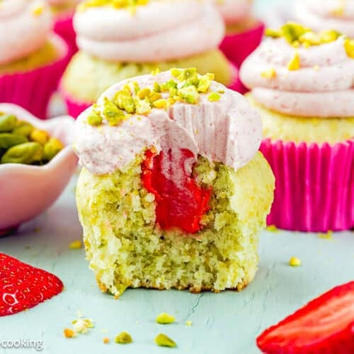 An Eggless Pistachio Cupcake with Strawberry Buttercream is shown with a bite taken out, revealing its strawberry filling. Whole cupcakes, a bowl of pistachios, and strawberries are arranged nearby.