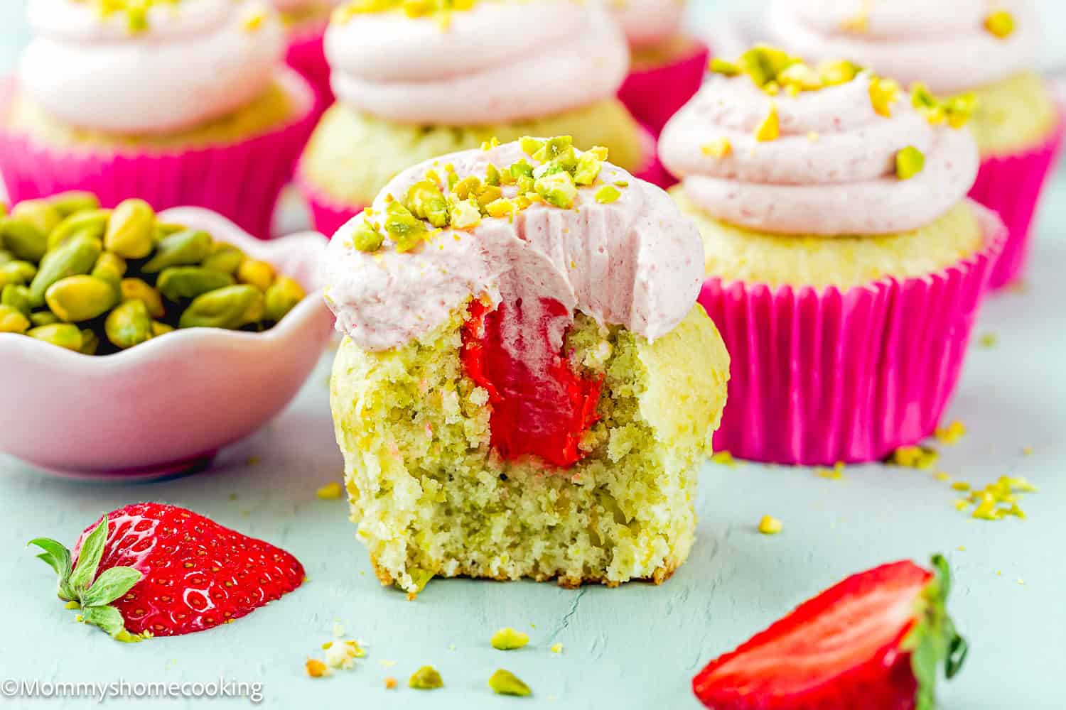 An Eggless Pistachio Cupcake with Strawberry Buttercream is shown with a bite taken out, revealing its strawberry filling. Whole cupcakes, a bowl of pistachios, and strawberries are arranged nearby.