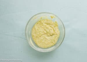 A glass bowl filled with thick, pale yellow batter for Eggless Pistachio Cupcakes with Strawberry Buttercream sits on a light blue surface.