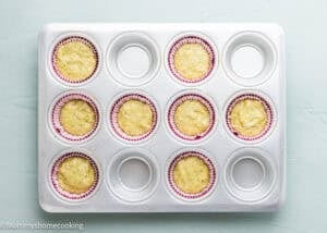 A metal muffin tin with six paper liners filled with batter for Eggless Pistachio Cupcakes with Strawberry Buttercream and six empty cups, placed on a light surface.