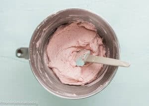 A mixing bowl filled with pink strawberry buttercream for Eggless Pistachio Cupcakes, with a spatula resting inside the bowl on a light blue surface.