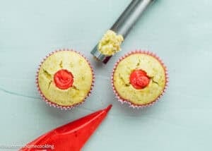 Eggless Pistachio Cupcakes with Strawberry Buttercream shown with red filling in the center, one being filled using a metal tool, and a red piping bag beside them on a light blue surface.