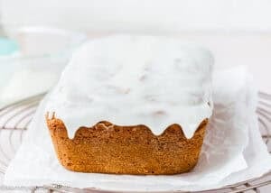 An Eggless Pound Cake Loaf topped with white icing sits on parchment paper, displayed on a wire cooling rack.