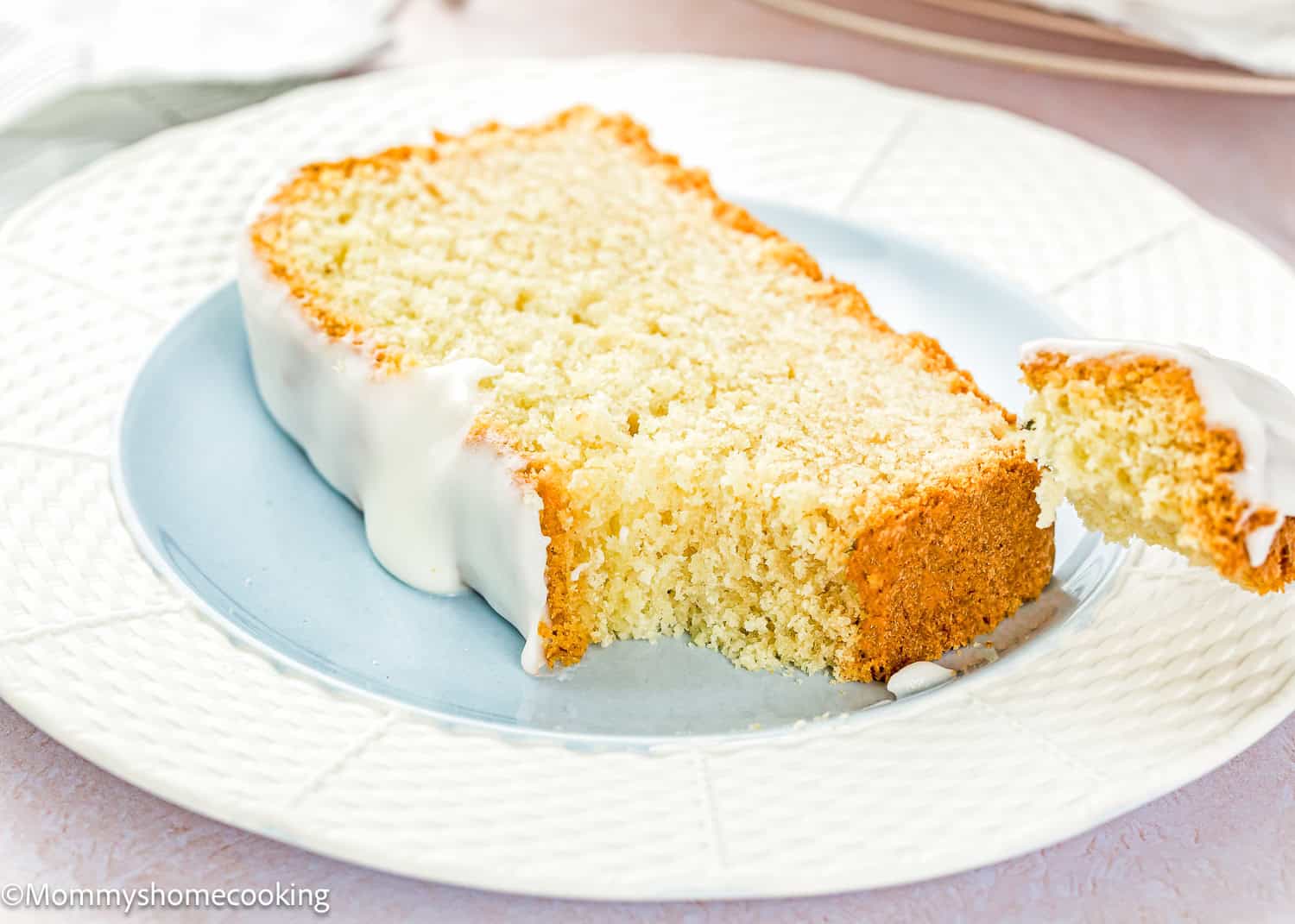 A slice of Eggless Pound Cake Loaf with white icing rests on a white plate, with a piece cut and lifted by a fork.