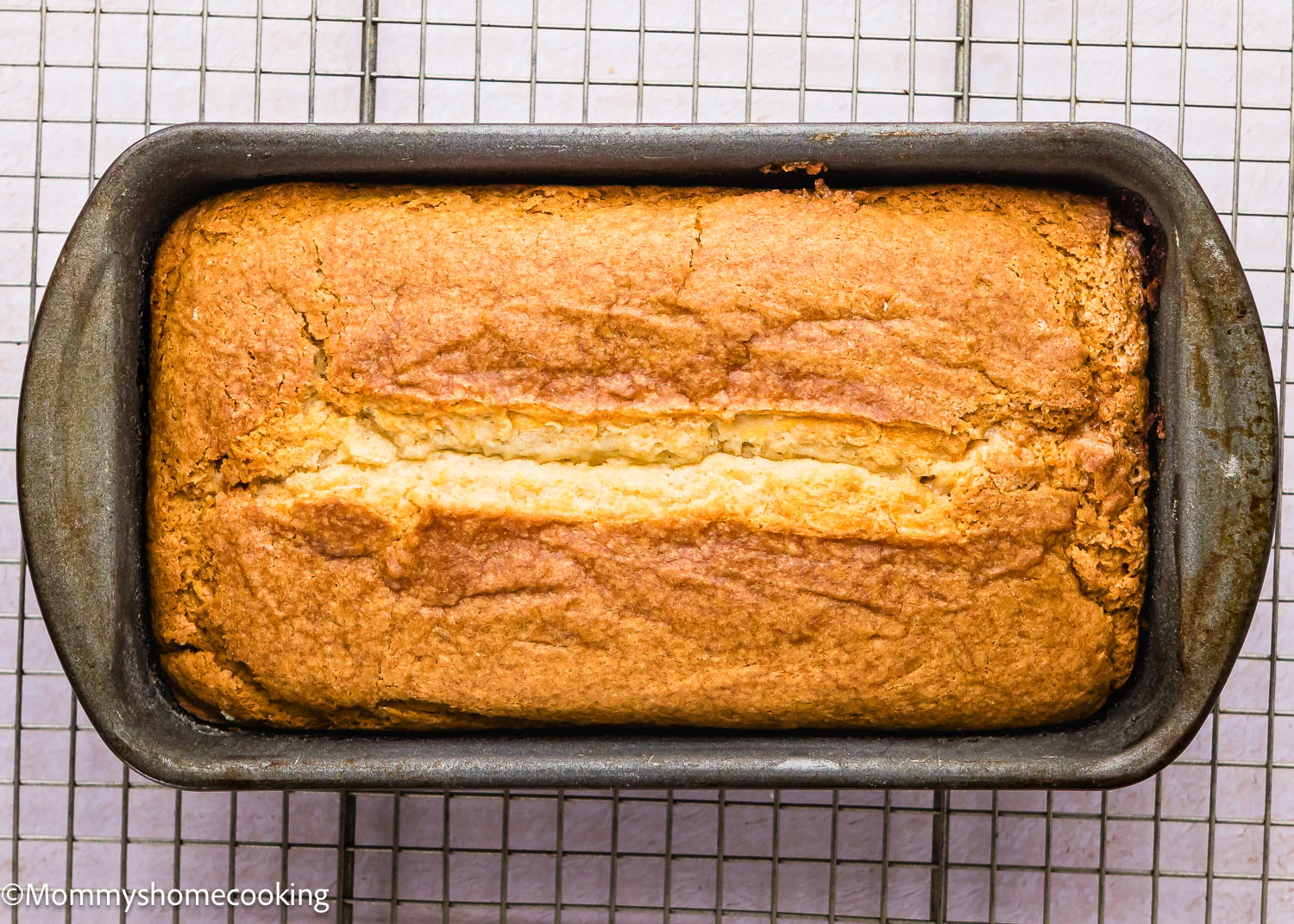 An Eggless Pound Cake Loaf with a cracked top sits in a metal pan on a wire cooling rack.