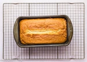 An Eggless Pound Cake Loaf in a metal pan sits on a wire cooling rack, its golden-brown crust inviting and warm.