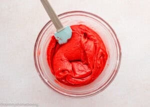 A glass bowl filled with bright red Eggless Strawberry Brownies batter being mixed with a light blue spatula on a light surface.