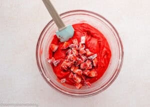 A glass bowl filled with red cake batter and chopped strawberries for eggless strawberry brownies, with a light blue spatula resting inside.