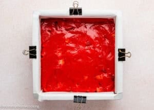 A parchment-lined square baking pan filled with smooth, bright red Eggless Strawberry Brownies batter, secured at the corners with black binder clips.