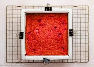 A square red Eggless Strawberry Brownie in a parchment-lined pan cools on a wire rack, secured with binder clips at the edges.