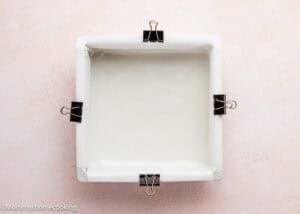 A square baking pan lined with parchment paper and secured with four black binder clips, ready for Eggless Strawberry Brownies, sits on a light pink surface.