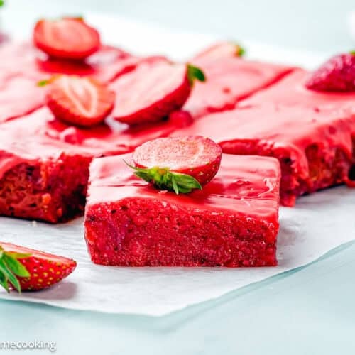 A batch of Eggless Strawberry Brownies with pink glaze, topped with halved fresh strawberries, sits on parchment paper. One piece is cut and placed in front.