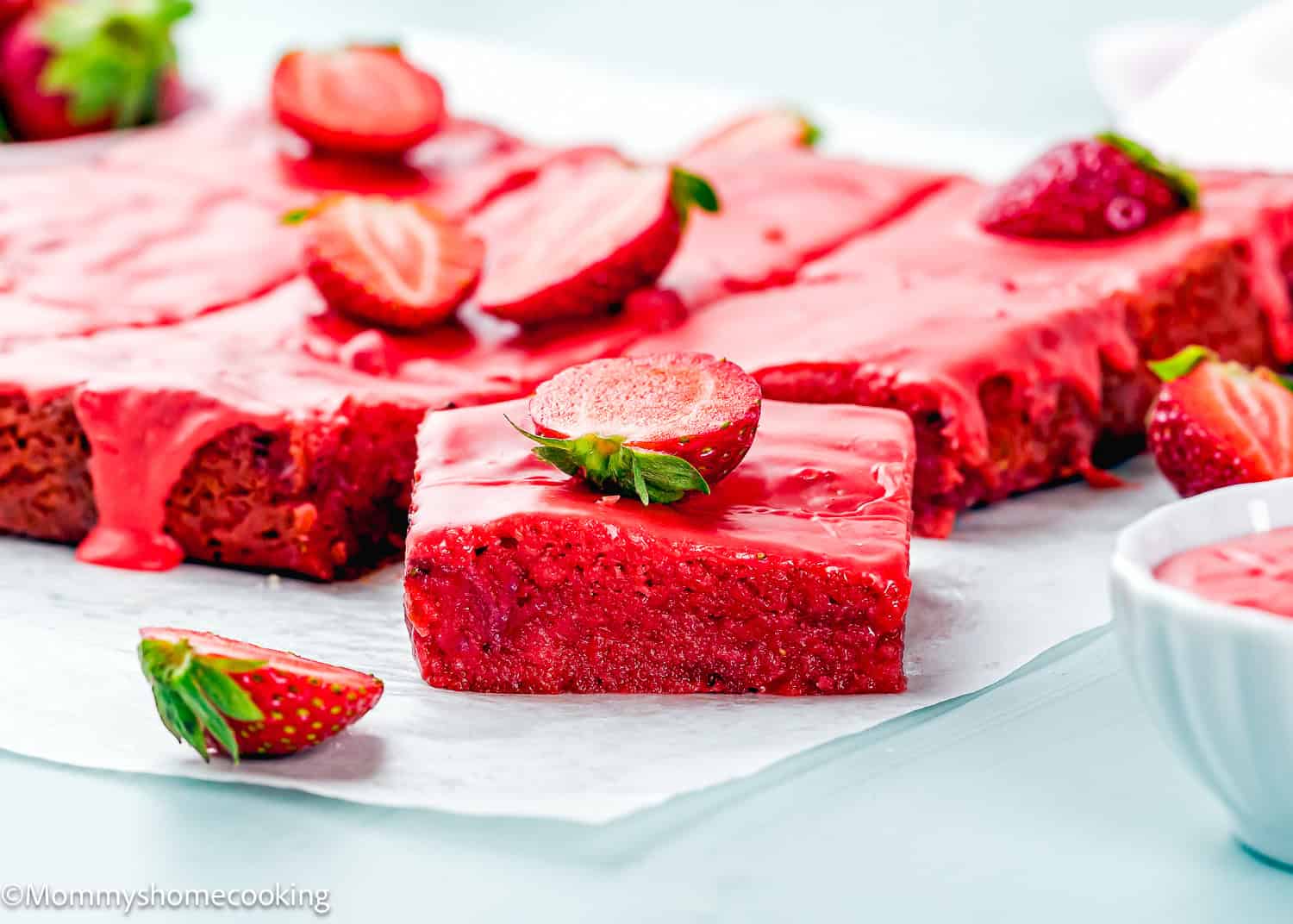A batch of Eggless Strawberry Brownies with pink glaze, topped with halved fresh strawberries, sits on parchment paper. One piece is cut and placed in front.