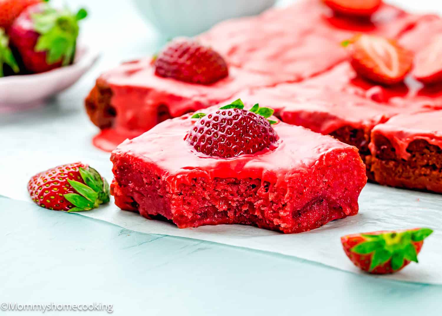 An eggless strawberry brownie topped with a halved strawberry sits on parchment paper, surrounded by fresh strawberries and more brownie pieces.