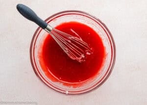 A glass bowl filled with red liquid mixture for eggless strawberry brownies and a whisk resting inside, placed on a light-colored surface.