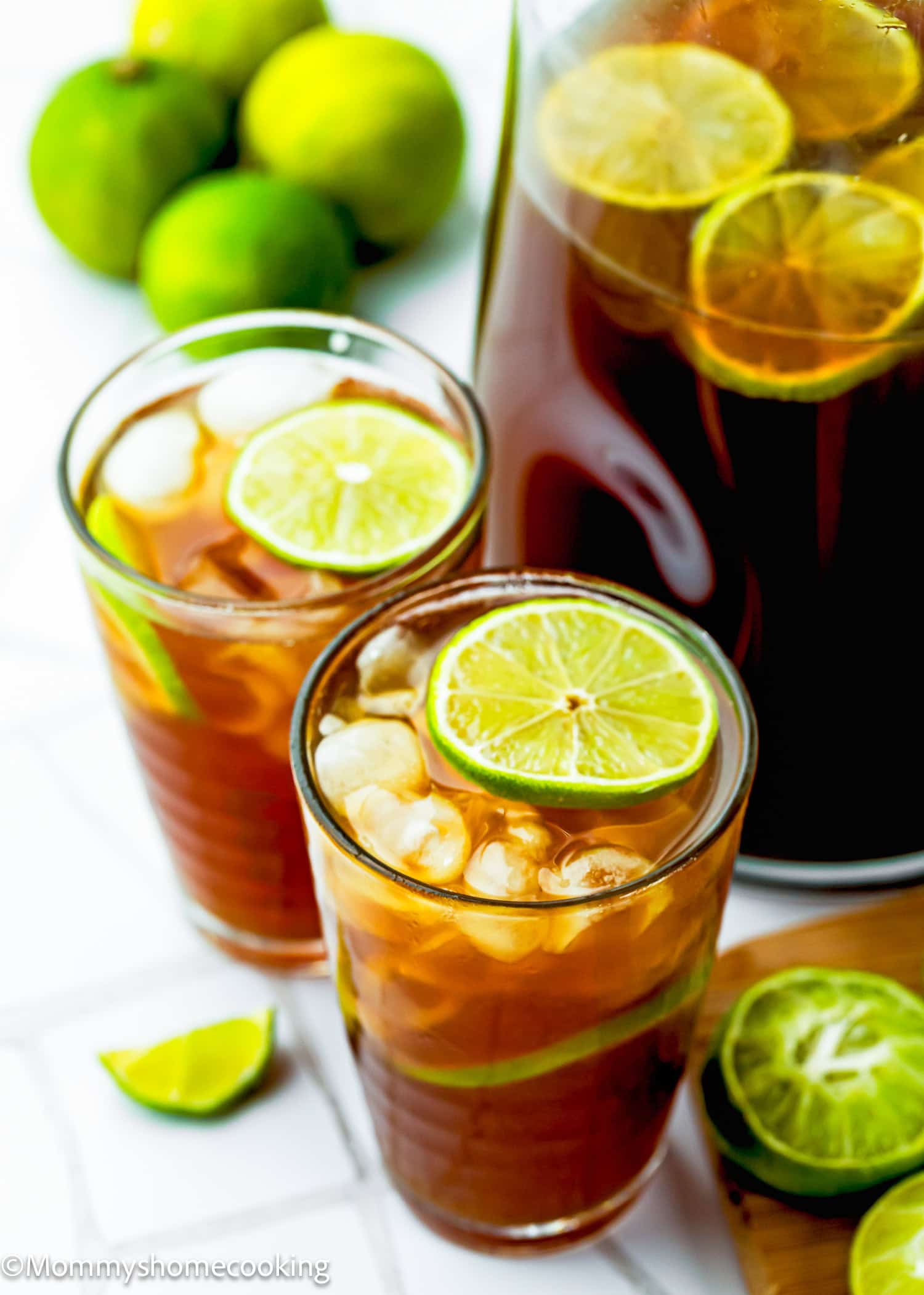 Two glasses of iced papelon con Limon with lime slices and ice, next to a pitcher of Papelón con Limón (Venezuelan Limeade) and fresh limes on a white surface.