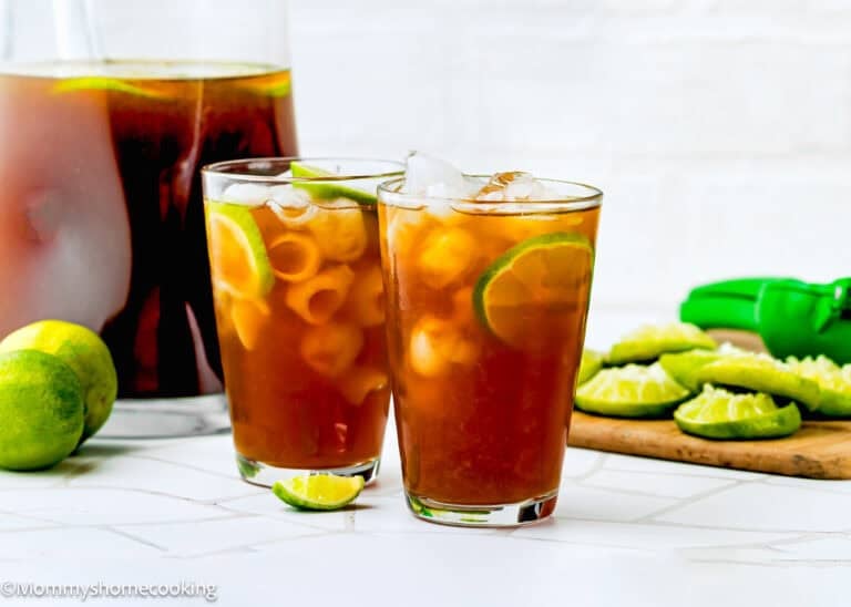Two glasses of iced papelon con limon with lime slices and ice cubes, a pitcher of papelon con limon, whole and sliced limes, a green citrus squeezer, on a white surface.