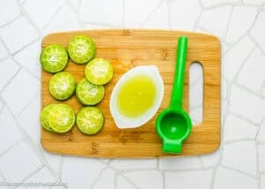 Sliced limes, a small bowl of lime juice, and a green lime squeezer sit on a wooden cutting board, ready to make Papelón con Limón (Venezuelan Limeade).