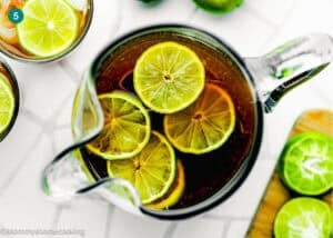 A glass pitcher filled with Papelón con Limón (Venezuelan Limeade) topped with lime slices, viewed from above on a white surface with limes nearby.