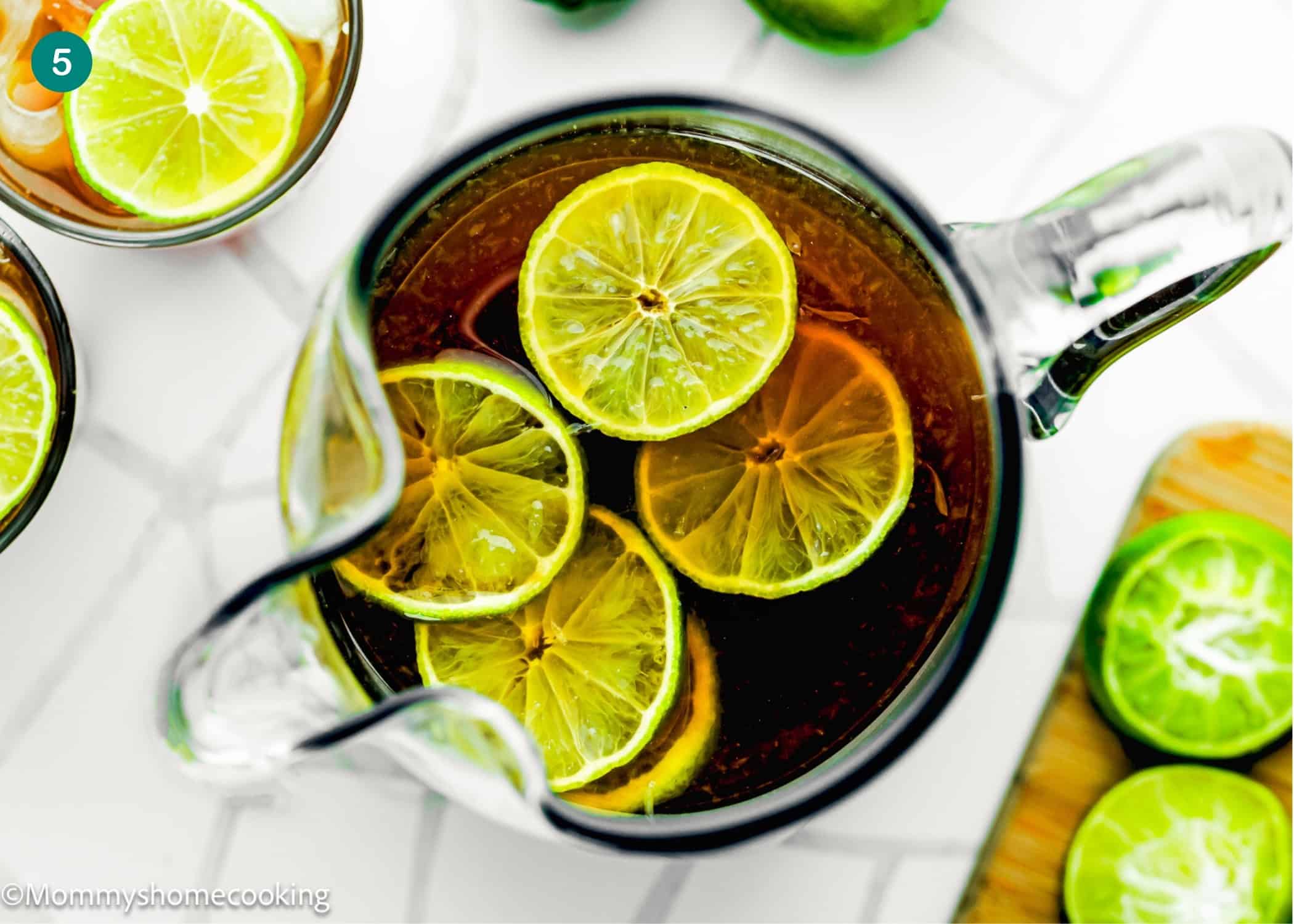 A glass pitcher filled with Papelón con Limón (Venezuelan Limeade) topped with lime slices, viewed from above on a white surface with limes nearby.