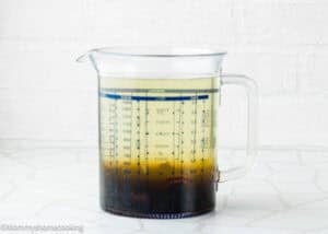 A clear measuring jug filled with Papelón con Limón (Venezuelan Limeade), its light yellow liquid and dark brown sediment at the bottom, sits on a white tiled surface.