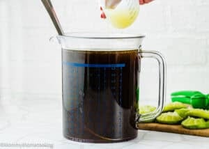 A person pours liquid from a small measuring cup into a large glass pitcher filled with Papelón con Limón (Venezuelan Limeade); sliced limes and a citrus squeezer are on the side.